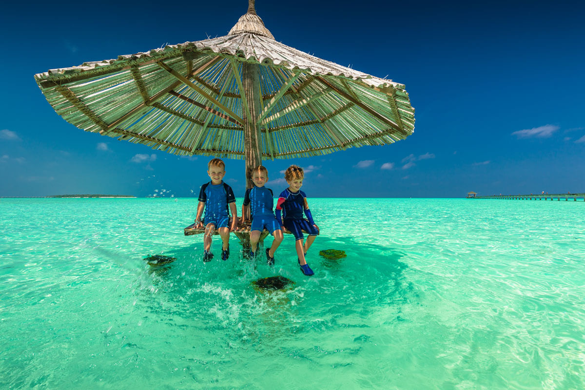 Photo de trois enfants assis sous un parasol, les pieds dans l’eau. 