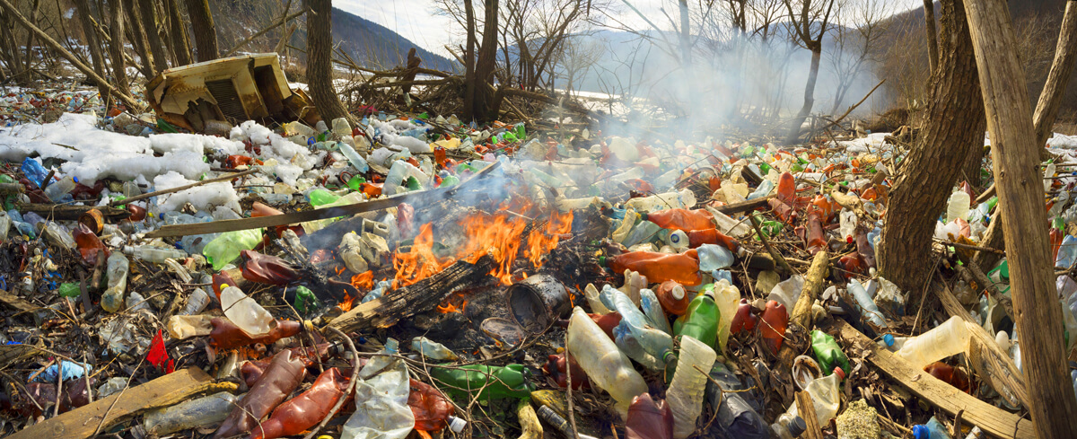 Photo d’un boisé rempli de déchets, en particulier de bouteilles en plastique. Au milieu des déchets, il y a du bois qui brûle. 